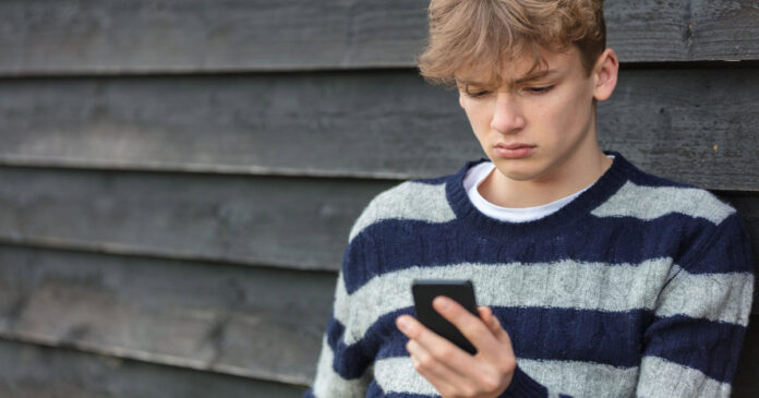Ein einsamer blonder Jugendlicher schaut besorgt auf sein Handy.
