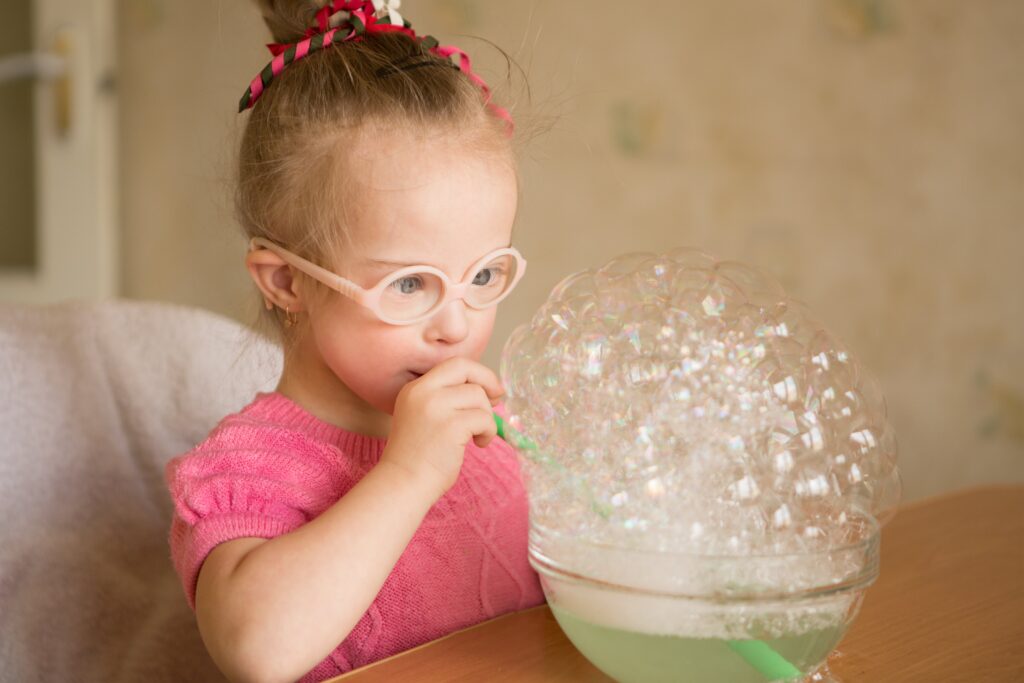 Kind mit Downsyndrom bläst in einen Strohhalm und macht Seifenblasen in einer Schale mit Seifenwasser.