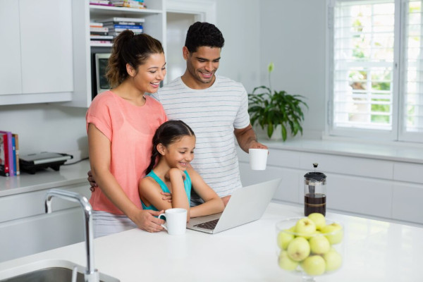 Zwei Eltern und ein Mädchen stehen vor dem Laptop in einer hellen Küche und lächeln.