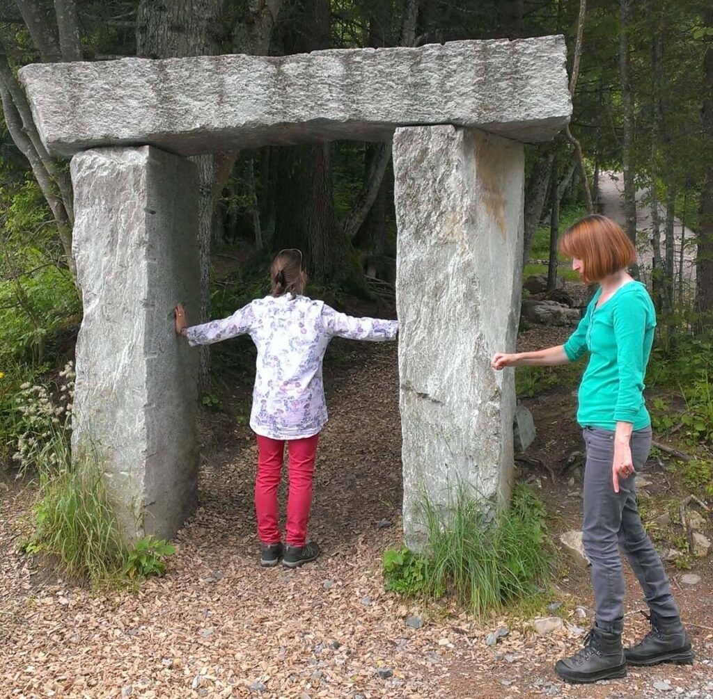 Frau steht in einem Steintor, eine weitere Frau steht mit einem Pendel in der Hand daneben.