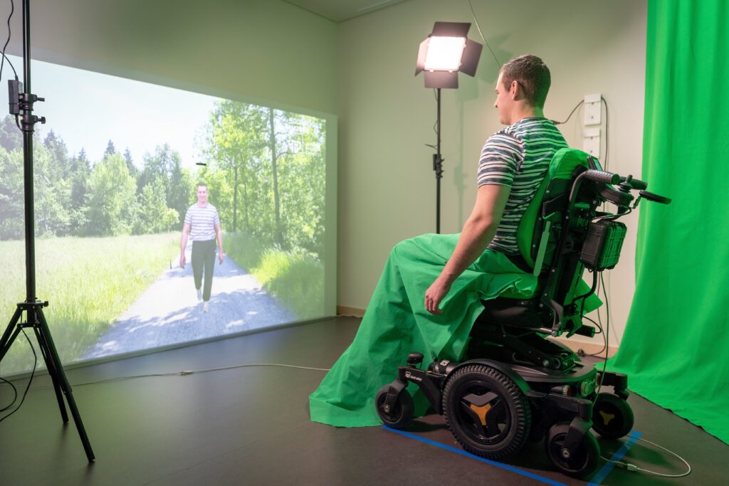 Ein Mann mit Querschnittslähmung sitzt in einem Rollstuhl und schaut auf die Leinwand, auf der er spazierend abgebildet ist.