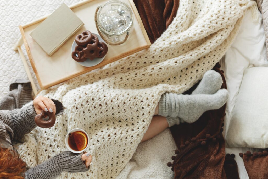 Frau liegt unter eine Decke gekuschelt und mit Pullover im Bett. Sie hat eine Tasse Tee in der Hand.