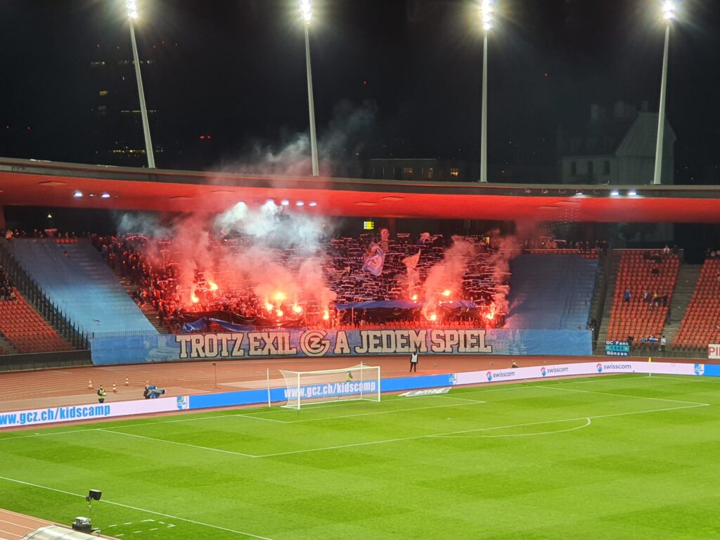 Das Feuerwerk in den Zuschauerreihen strahlt in Rot und produziert viel Rauch. Die Fans jubeln.