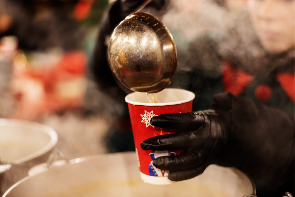 Eine Person mit schwarzen Handschuhen giesst Glühwein in einen Pappbecher.