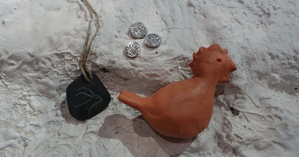 Ein Anhänger, drei keltische Münzen und ein aus Ton geformtes Huhn liegen auf einem Stein. Solche und ähnliche Basteleien kannst du im Workshop gestalten.