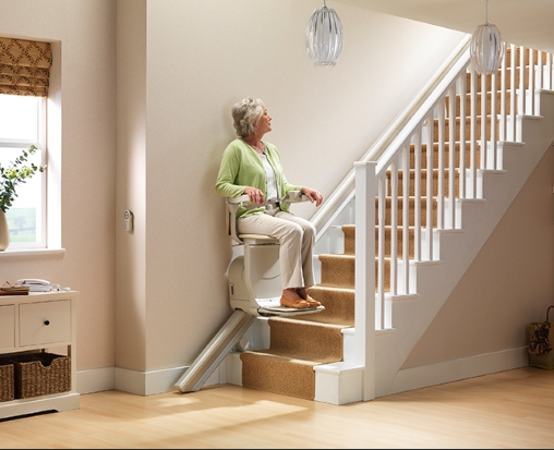 Eine ältere Frau sitzt auf einem Treppenlift am Fusse einer Treppe in einem Haus