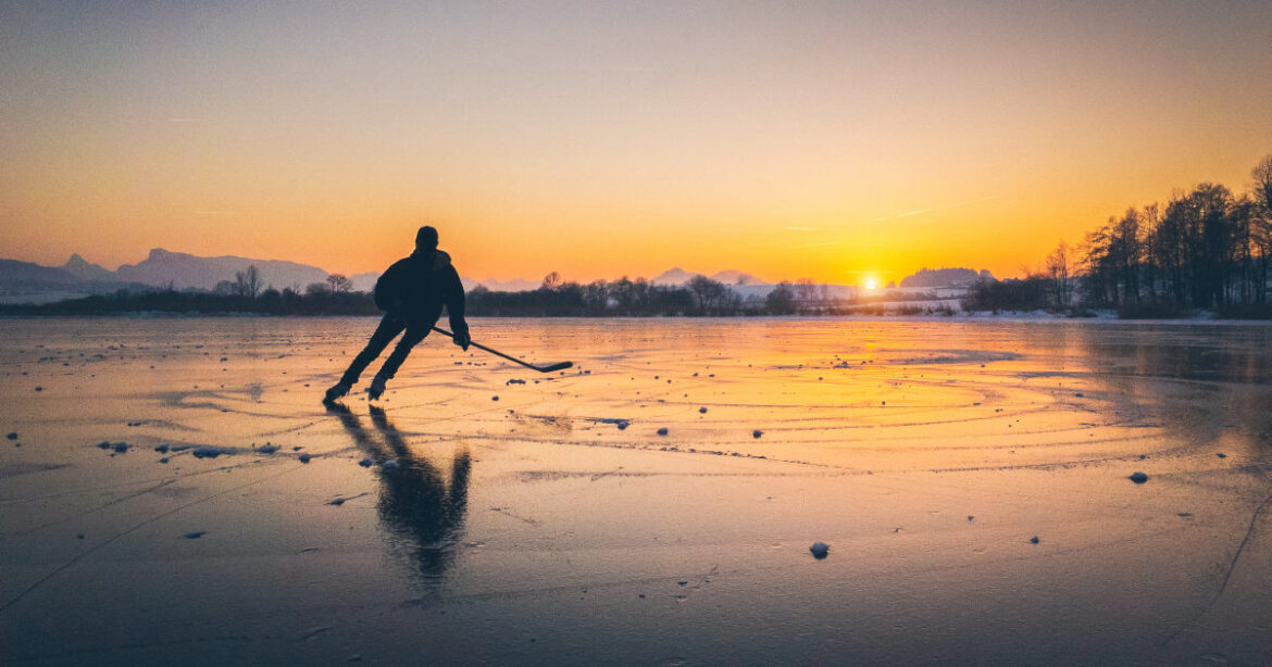 Mann mit Schläger und Schlittschuhen auf zugefrorenen See beim Sonnenuntergang