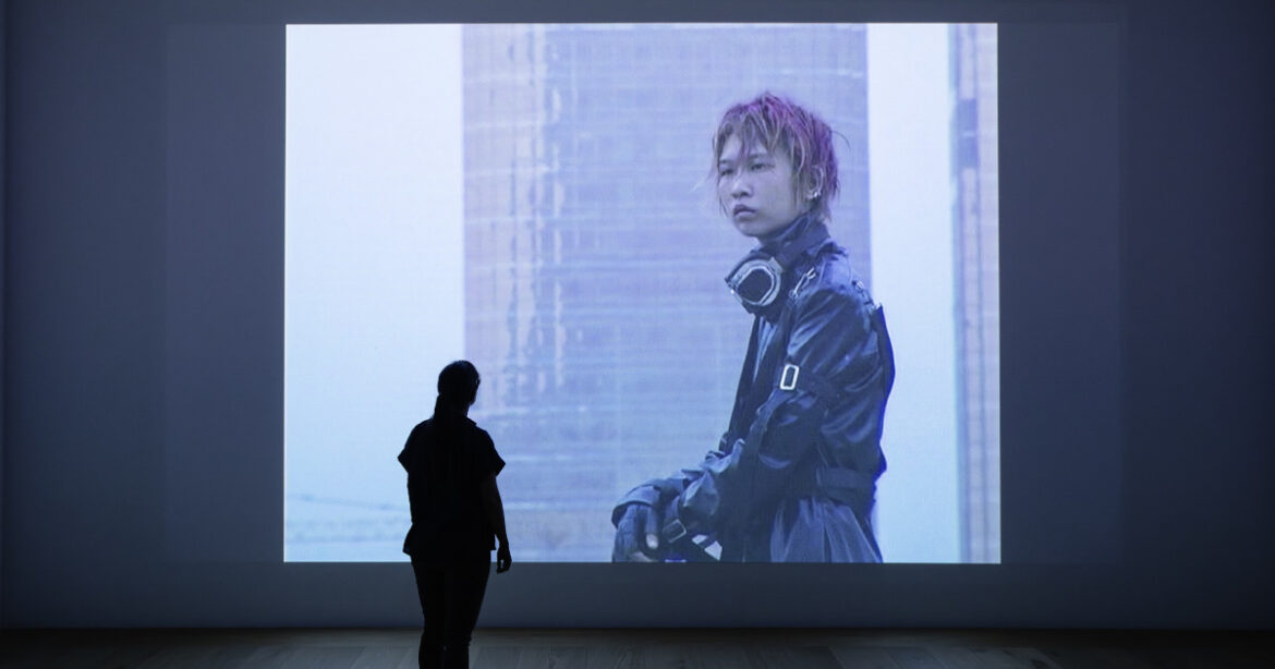 Frau steht vor Leinwand, darauf Videoprojektion von junger Chinesin im Kostüm