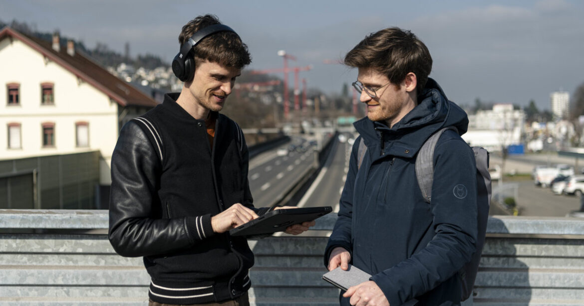 Zwei junge Männer stehen auf der Autobahnbrücke, die linke Person hat Kopfhörer und ein Tablet