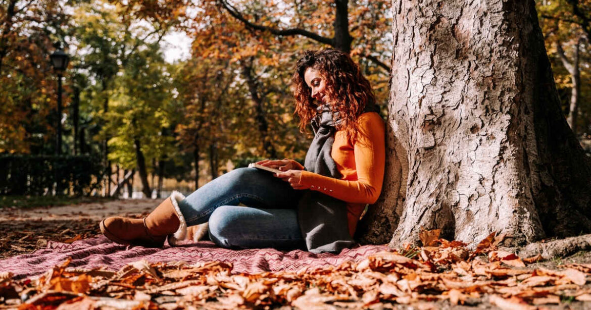 Frau liest auf dem Waldboden sitzend und an einen Baum gelehnt ein Buch im Herbst