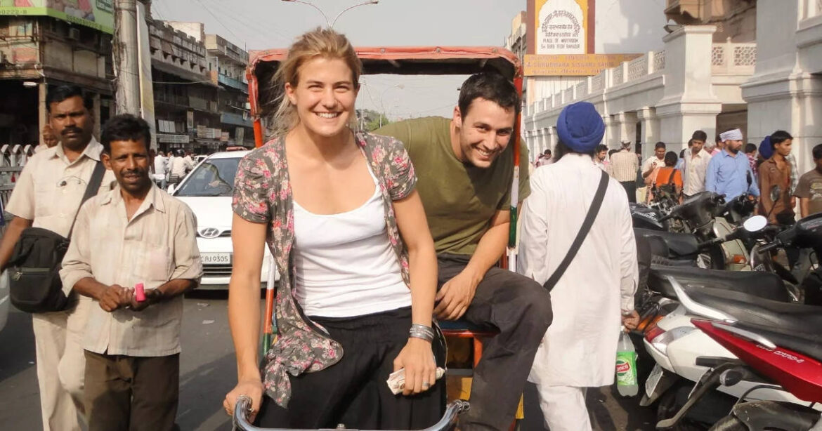 Daniela Widmer und David Och sitzen auf einer Rikscha und lachen auf einer Strasse in Indien. Leute gehen an ihnen vorbei