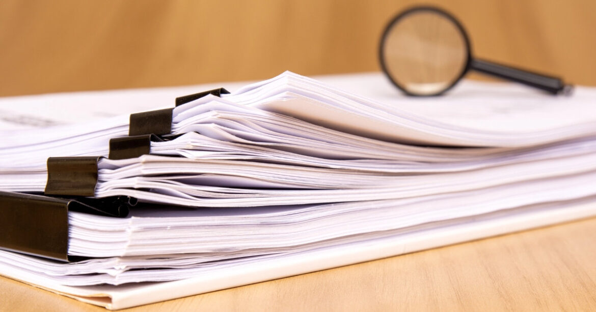 ein Stapel an Dokumenten liegt auf einem Tisch. Darauf eine Lupe