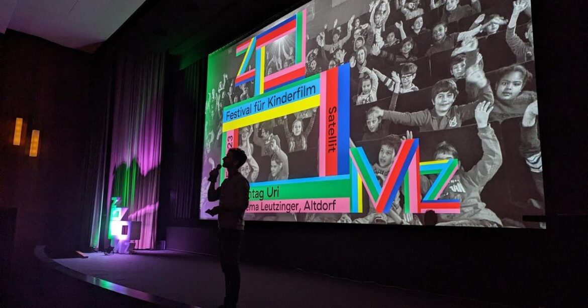 Auf der Leinwand eines Kinos ist das Logo von Zoomz zu sehen, davor die Silhouette eines Mannes mit einem Mikrofon.