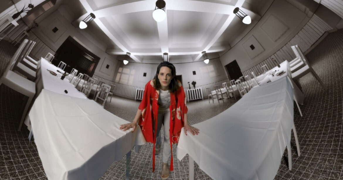 Eine Frau blickt in die Kamera, die Hände auf zwei Tischen aufgestützt. Dies im Rahmen des VR-Erlebnisses Zauberberg: Expedition in die Gegenwart