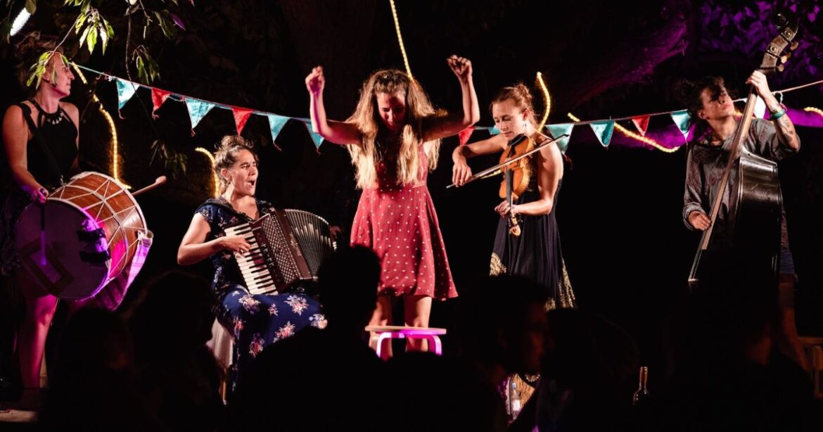 Haimos singen, tanzen und spielen ihre Instrumente auf einer Bühne. Der Kontrast zwischen dem dunklen Hintergrund und den farbigen Kleidern der Frauen fällt auf.