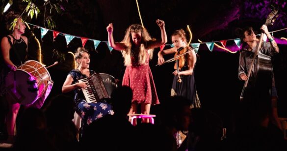 Haimos singen, tanzen und spielen ihre Instrumente auf einer Bühne. Der Kontrast zwischen dem dunklen Hintergrund und den farbigen Kleidern der Frauen fällt auf.