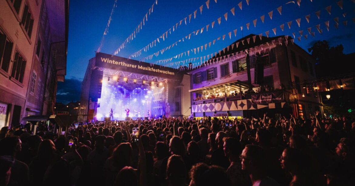 Auf der Steinberggasse in Winterthur steigen die Musikfestwochen - auf der Bühne sind MusikerInnen zu sehen sowie grelle Lichter, während die Gasse selbst voller Menschen ist.