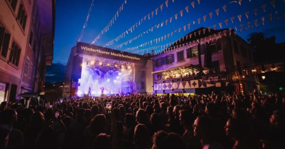 Auf der Steinberggasse in Winterthur steigen die Musikfestwochen - auf der Bühne sind MusikerInnen zu sehen sowie grelle Lichter, während die Gasse selbst voller Menschen ist.