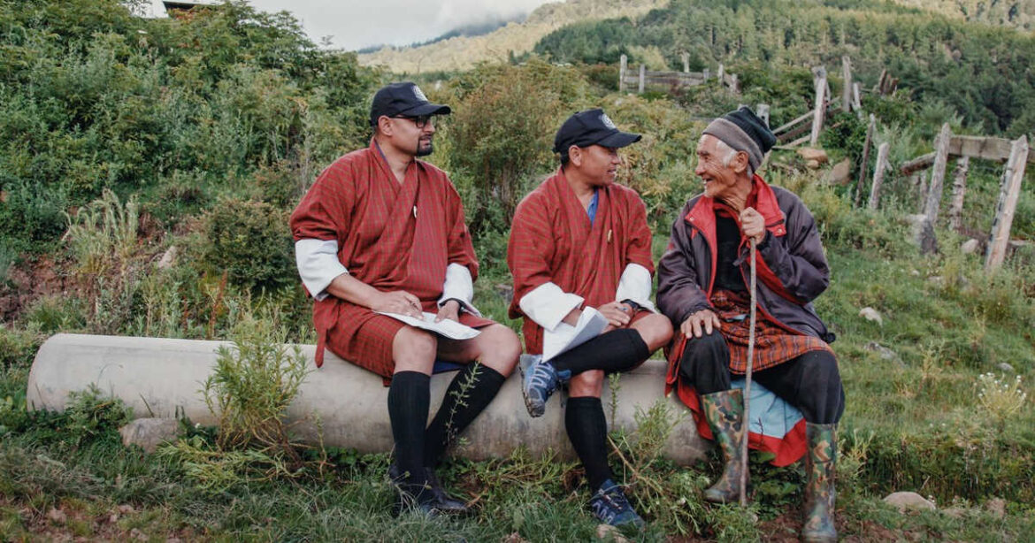 Die Glücksagenten Amber und Guna haken nach bei einer älteren Person, sie sitzen auf einem Baumstamm im Dokumentarfilm Agent of Happiness aus Bhutan