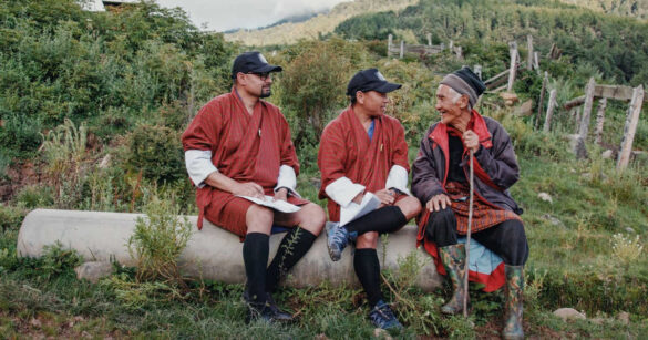 Die Glücksagenten Amber und Guna haken nach bei einer älteren Person, sie sitzen auf einem Baumstamm im Dokumentarfilm Agent of Happiness aus Bhutan