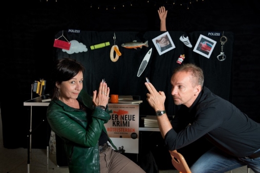 Judith Stadlin und Michael van Orsouw setzen auf Stühlen, dazu haben sie die Hände zu Pistolen gefaltet