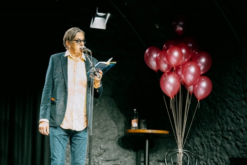 Pedro Lenz steht auf der Bühne des Theaters Burgbachkeller und liest aus einem Buch am Literaturfestival Höhenflug