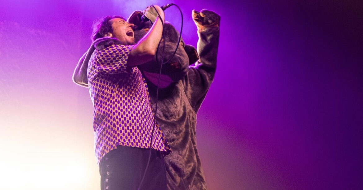 The Wombats-Frontmann Matthew Murphy mit einem kostümierten Wombat auf der Bühne. Er singt in ein Mikrofon